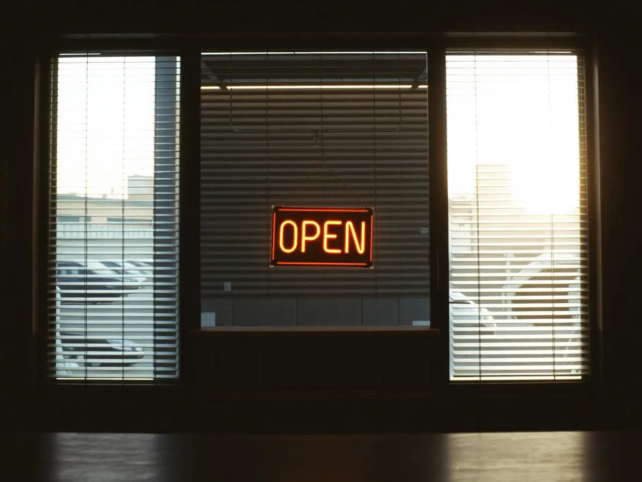 Empty small-shop interior at dawn with an OPEN sign hanging in the window and soft light through blinds.